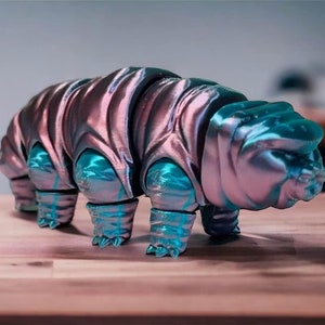 Puede incluir: Una figurita impresa en 3D de un tardígrado, también conocido como oso de agua, en un esquema de color rosa metálico y turquesa. La figurita está en posición de caminar con su cuerpo segmentado y ocho patas visibles.