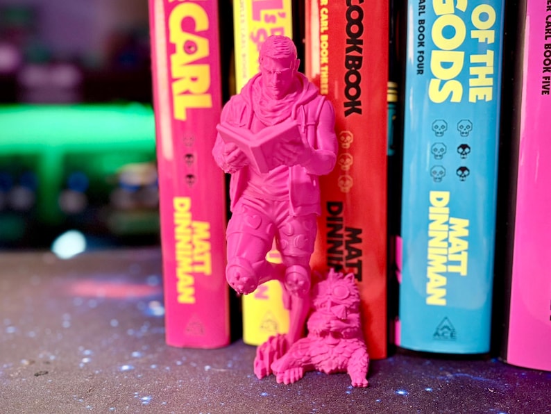 May include: A vibrant pink figurine of a person reading a book, with a small animal at its feet, stands before a row of colorful books. The books have titles and author names in yellow, red, and blue.