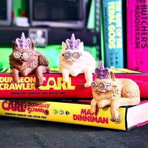 May include: Three cat figurines wearing glasses and purple crowns rest on top of books. The books are titled "Dungeon Crawler Carl" and "Carl Doomsday Scenario". Other books are visible in the background.