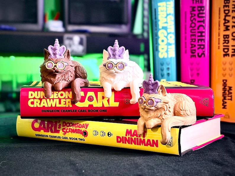 K&ouml;nnte beinhalten: Drei Katzenfiguren mit Brille und lila Kronen liegen auf B&uuml;chern. Die B&uuml;cher tragen die Titel "Dungeon Crawler Carl" und "Carl Doomsday Scenario". Weitere B&uuml;cher sind im Hintergrund zu sehen.