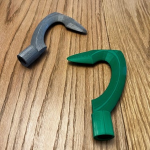 May include: Two 3D printed hooks, one gray and one green, are on a wooden surface. The hooks are curved and have a rounded end.