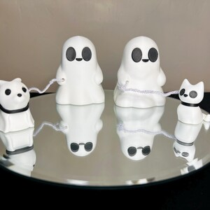 Pode incluir: Tr&ecirc;s figurinhas de fantasmas brancas de cer&acirc;mica com olhos e bocas pretos. Dois dos fantasmas seguram cordas brancas, e um fantasma segura uma corda branca amarrada a uma figurinha de cachorro branca de cer&acirc;mica com olhos pretos e uma coleira preta. As figurinhas est&atilde;o sobre uma superf&iacute;cie de espelho prateado.