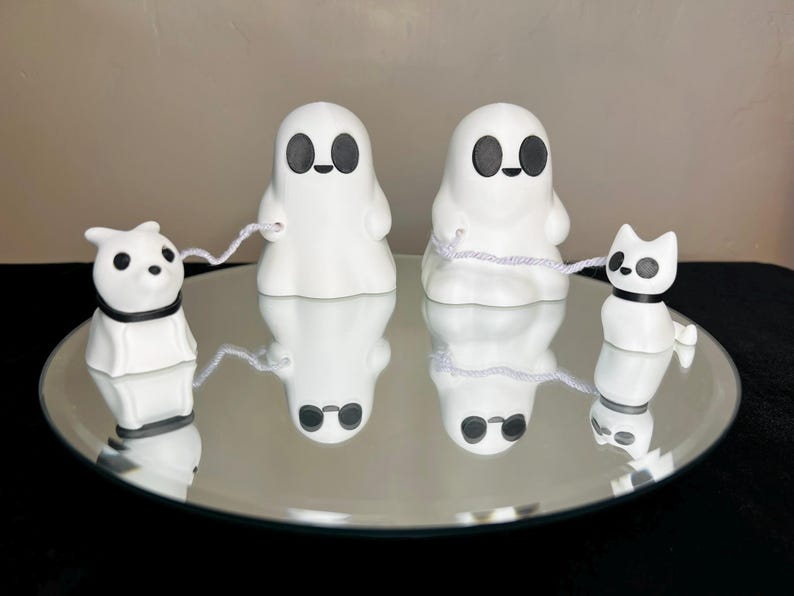 Pode incluir: Tr&ecirc;s figurinhas de fantasmas brancas de cer&acirc;mica com olhos e bocas pretos. Dois dos fantasmas seguram cordas brancas, e um fantasma segura uma corda branca amarrada a uma figurinha de cachorro branca de cer&acirc;mica com olhos pretos e uma coleira preta. As figurinhas est&atilde;o sobre uma superf&iacute;cie de espelho prateado.