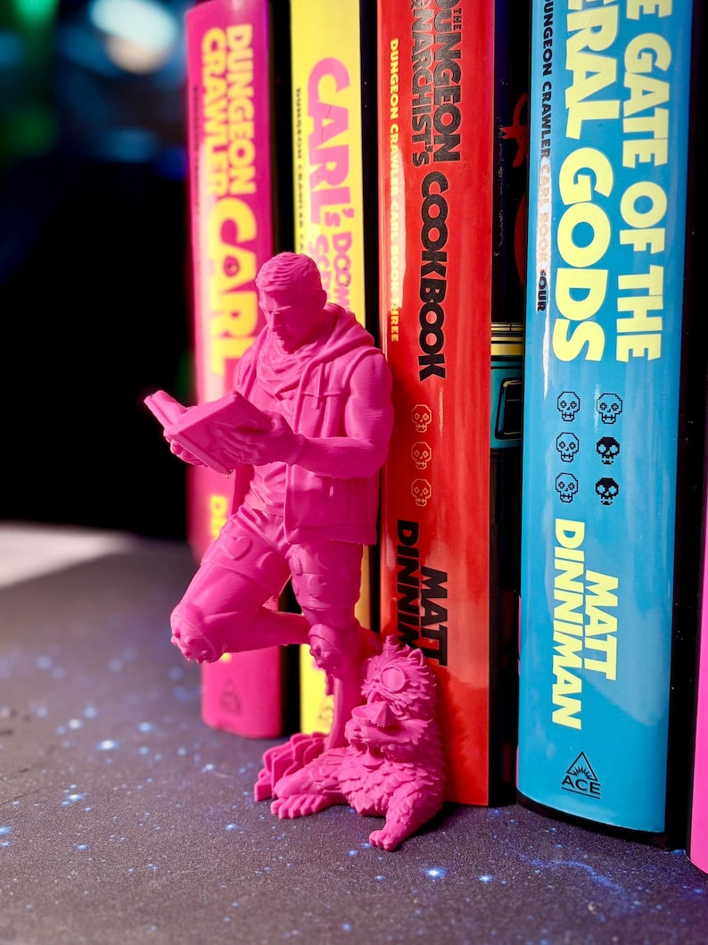 May include: A vibrant pink figurine of a person reading a book, with a cat figurine at their feet, positioned in front of a row of colorful books. The books have titles like "Dungeon Crawler" and "The Gate of the Eternal Gods."