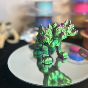 May include: A three-headed green and purple dog figurine with a shiny finish. The figurine is standing on a round, silver-colored mirror.