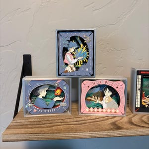 Puede incluir: Varias obras de arte enmarcadas con personajes de anime. Las obras de arte están en cajas transparentes y se exhiben en una estantería de madera. Las obras de arte tienen una variedad de colores, incluyendo azul, rosa y amarillo. Algunas obras de arte tienen texto, incluyendo el nombre "SOPHIE".