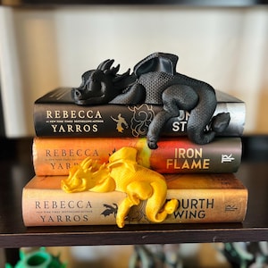 May include: A black dragon figurine resting on a book titled Onyx Storm and a yellow dragon figurine resting on a book titled Fourth Wing. The books are stacked vertically, with Iron Flame in the middle, all authored by Rebecca Yarros.