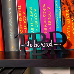 May include: A black wooden TBR (To Be Read) book stand with the words "to be read" in a cursive font. The stand is placed in front of a row of colorful books on a shelf.