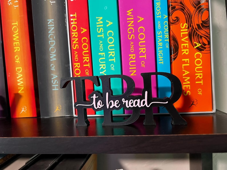 May include: A black wooden TBR (To Be Read) book stand with the words "to be read" in a cursive font. The stand is placed in front of a row of colorful books on a shelf.