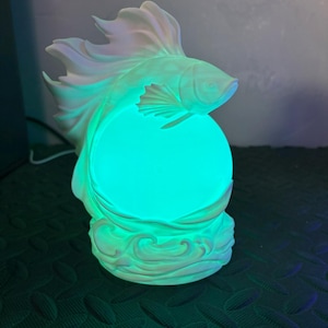 Puede incluir: Una taza de cerámica blanca con un asa negra y el texto "But first, coffee" en un estilo manuscrito. La taza está sobre una superficie blanca. La taza mide aproximadamente 10 cm de alto.