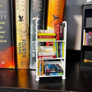 Peut inclure: Une petite étagère blanche à roulettes, remplie de livres colorés. L'étagère est placée devant une rangée de livres plus grands. Les livres présentent diverses couleurs et titres, créant un affichage visuellement attrayant. La scène évoque un coin lecture confortable.