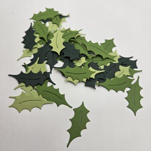 以下が含まれることがあります： さまざまな緑色の色合いのヒイラギの葉の切り抜き。 葉は詳細なとげのあるエッジを持ち、白い表面に散らばっています。 クラフトやホリデーデコレーションに最適で、季節のテーマを連想させます。
