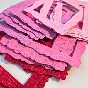 Könnte beinhalten: Ein Stapel dekorativer Papierrahmen in Rosa- und Rottönen. Die Rahmen haben verzierte, wellenförmige Kanten und detaillierte geprägte Muster. Die Rahmen sind gestapelt, wobei sich einige überlappen, wodurch ein geschichteter Effekt entsteht.