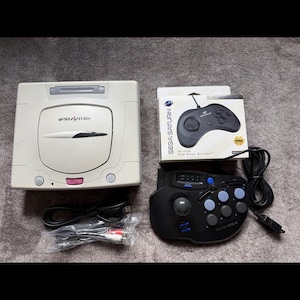 May include: A Sega Saturn video game console, a black controller, and a controller in its original box. The console is off-white with a disc tray and the Sega Saturn logo. The controllers are black with blue buttons.