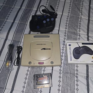 May include: A beige Sega Saturn game console, a black arcade-style joystick controller, a boxed black game controller, a video cable, and a game cartridge. The console features the Sega Saturn logo on the top lid and a purple power button.