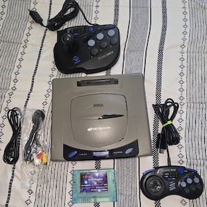 May include: A grey Sega Saturn game console with a black Eclipse Stick arcade controller, a black Eclipse Pad controller, a blue translucent SS HDloader game cartridge, and two black power and AV cables with yellow, white, and red connectors.