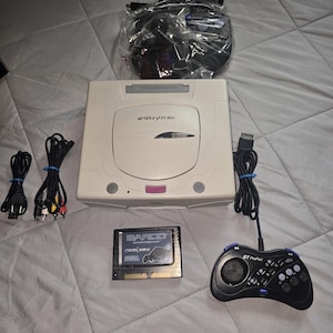 May include: A beige Sega Saturn video game console with a disc drive lid, a black ST ProPad controller, a SAROO flash cartridge, a power cable, and an AV cable. An additional black controller is included, sealed in a clear plastic bag.