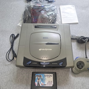 May include: A gray Sega Saturn video game console with a matching controller, game cartridge, and power cord. The console has the Sega Saturn logo and buttons for power and access. The game cartridge has a colorful anime-style graphic.