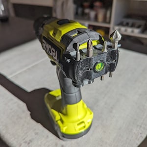 Puede incluir: Un taladro inalámbrico amarillo y negro con un soporte de brocas negro y varias brocas de taladro adjuntas. El taladro tiene un indicador de nivel verde y la palabra "RYOBI" es visible en el lateral.