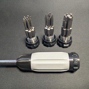 Op de afbeelding: Een multifunctionele schroevendraaier met een lichtgrijze en donkergrijze handgreep en een metalen schacht, vergezeld van drie ronde opbergers die elk een cluster van diverse metalen schroevendraaierbits in verticale stand bevatten.