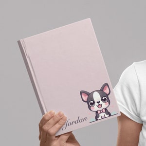 Op de afbeelding: Een roze gepersonaliseerd notitieboek met een cartoon Franse bulldog die een strikje draagt. Het notitieboek heeft de naam "Jordan" erop.