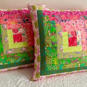 May include: Two square cushions with a patchwork design. The cushions feature a vibrant mix of pink, green, and floral patterns. The patchwork includes squares and strips of fabric in various shades and prints, creating a colourful and textured look.
