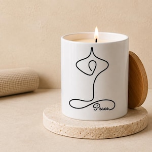 May include: A white ceramic candle jar with a lit wick and a wooden lid. The front features a black line art illustration of a person in a seated yoga pose with the word Peace written in a cursive script at the base of the design.