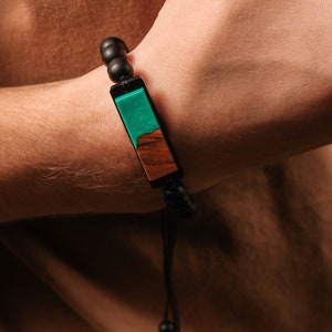 May include: A black beaded bracelet with a rectangular wooden charm. The charm is made of two different types of wood, one green and one brown. The bracelet has a black cord with black beads at the end.