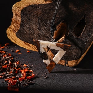 May include: A triangular pendant necklace with a white and brown resin design. The pendant is hanging from a black cord and is resting on a piece of dark wood.