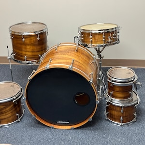 Puede incluir: Un set de batería de cinco piezas con un acabado en madera marrón. El set incluye un bombo, una caja, dos toms y un tom de piso. El bombo tiene un logotipo negro de Aquarian en la parte delantera.