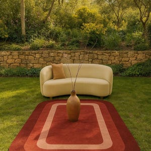 May include: An outdoor scene features a beige sofa with a brown pillow, a rust-colored rug with a lighter border, and a brown vase with branches. The setting is a grassy area with a stone wall and trees in the background.