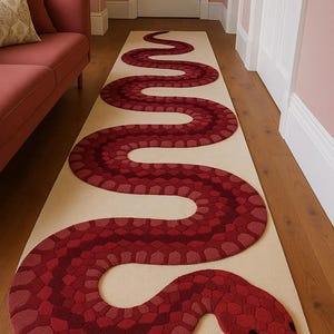 Puede incluir: Una alfombra larga y beige con un diseño de serpiente roja. El cuerpo de la serpiente está formado por varios tonos de rojo, creando una apariencia texturizada. La cabeza de la serpiente presenta ojos negros y una lengua roja. La alfombra está sobre un suelo de madera.