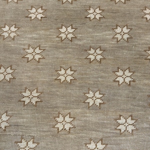 May include: A close-up of a beige rug with a repeating pattern of star-shaped floral designs in a lighter beige color. The rug has a textured surface and a border at the top.