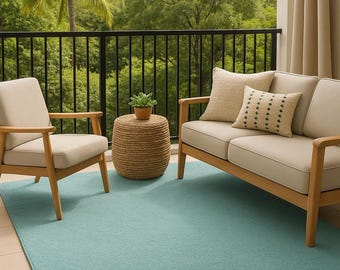 May include: An outdoor living space featuring a light blue rug, a two-seater sofa, and an armchair with beige cushions and wooden frames. A small woven side table holds a potted plant. The background includes a black railing and lush greenery.