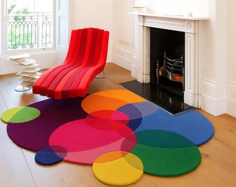 May include: A modern living room scene featuring a red chaise lounge and a colorful, abstract rug. The rug has overlapping circles in shades of purple, pink, orange, blue, and green. A white fireplace and window are in the background.