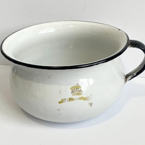 May include: White enamel chamber pot with a black rim and handle. The pot has a rounded shape and a small handle on one side. There is some wear and tear on the surface, with a faded yellow design.