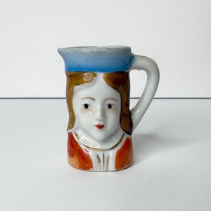 May include: A small ceramic pitcher shaped like a human head with brown hair, a blue headband, and a red garment. The pitcher features a white face with painted facial features, a handle on the right side, and a spout at the top rim for pouring.