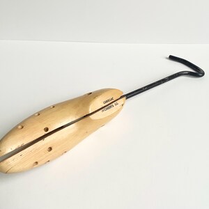 May include: A wooden shoe stretcher with a black metal handle. The shoe stretcher is light brown and has several small holes. The text "Cadillac Women's LG" is printed on the side.