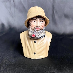 May include: A ceramic figurine of a weathered fisherman with a beige hat and coat. The figurine has a detailed face with a white beard and is smoking a pipe with a red bowl. The bust is a vintage nautical collectible.