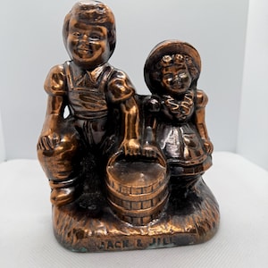 May include: A copper-colored figurine of Jack and Jill, with Jack holding a bucket and Jill standing beside him. The figurine is on a base with the words "Jack & Jill" inscribed on it.