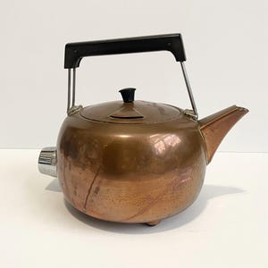 May include: A vintage copper electric kettle with a black handle and lid knob. The kettle has a spout and a chrome-colored heating element on the side. The kettle is round and sits on small feet.