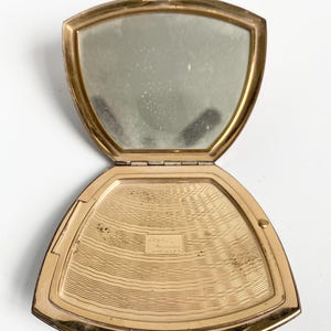 May include: Open vintage gold-toned compact mirror with a hinged lid. The interior features a mirror on one side and a textured, gold-toned surface on the other. The compact is open, revealing the mirror and the powder compartment.
