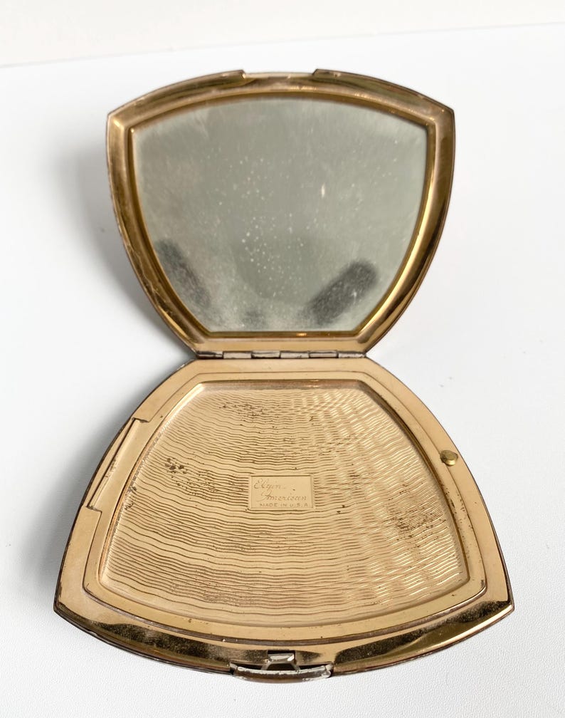 May include: Open vintage gold-toned compact mirror with a hinged lid. The interior features a mirror on one side and a textured, gold-toned surface on the other. The compact is open, revealing the mirror and the powder compartment.