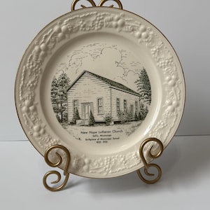 May include: A white ceramic plate with a raised border of grapes and leaves. The center of the plate features a black and white line drawing of a church building with the text "New Hope Lutheran Church, Saltillo, Mississippi, Birthplace of Mississippi Synod, 1855-1955".