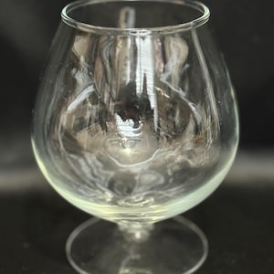 May include: A clear, empty brandy snifter glass with a rounded bowl and a short stem. The glass is made of transparent material, allowing light to pass through. The glass sits on a circular base, and the rim is smooth.