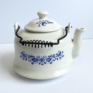 May include: A white ceramic teapot with a lid and a black wire handle. The teapot features blue floral patterns and a spout. The handle is made of black wire. The teapot is designed for brewing tea.