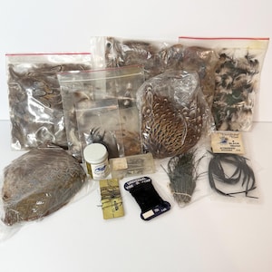 May include: A collection of fly tying materials including various bags of brown and white feathers, a small jar of cement, a spool of black thread, a bundle of dark fibers, a pack of black stripped goose quills, and a small box of metal hooks.