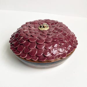 May include: A ceramic decorative bowl with a lid. The lid is covered in burgundy petal-shaped designs, with a small bee-like figure on top. The bowl has a dark blue and brown base.