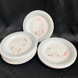May include: A set of white ceramic plates with a light blue border and floral design. The plates feature a delicate pattern of pink and white flowers with green leaves. The plates are stacked and arranged on a black surface.