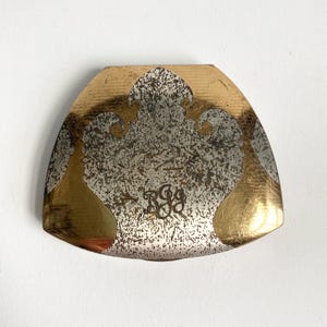 May include: A gold and silver-toned metal object with a curved, shield-like shape. The top and edges are gold, while the center features a textured silver design with the initials "RGG" in a decorative script. The object has a vintage aesthetic.